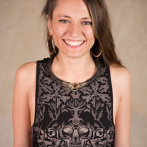 May include: Black tank top with a circuit board and skull design. The tank top has a round neckline and a sleeveless design. The model is wearing a black necklace with a circular pendant and gold hoop earrings.