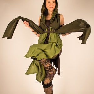 May include: A woman wearing a green and brown hooded cloak with a long flowing scarf. She is standing with one leg bent and her arms outstretched.