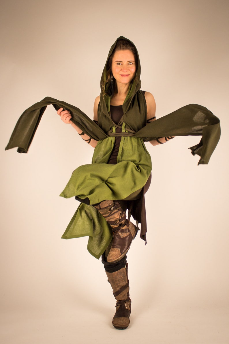 May include: A woman wearing a green and brown hooded cloak with a long flowing scarf. She is standing with one leg bent and her arms outstretched.