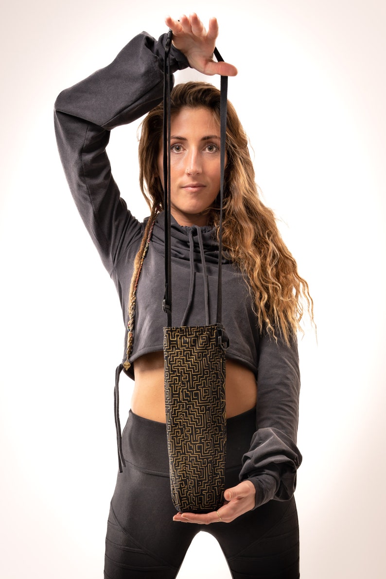 May include: A black and gold patterned water bottle holder with a black strap. The holder is held up by a person. The holder has a geometric design in gold on a black background. The strap is black and appears to be adjustable.