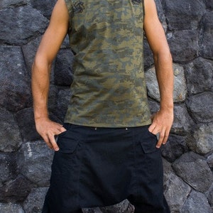 Op de afbeelding: Een man draagt een mouwloos camouflage shirt met zwarte accenten en een zwarte cargo broek. Hij staat voor een stenen muur.