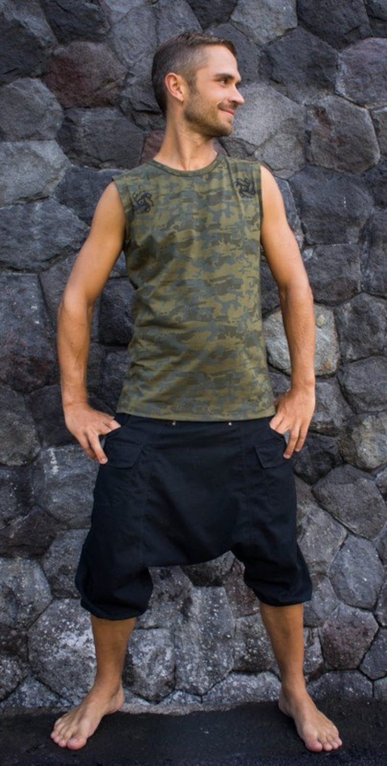 Op de afbeelding: Een man draagt een mouwloos camouflage shirt met zwarte accenten en een zwarte cargo broek. Hij staat voor een stenen muur.