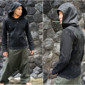 May include: Black hooded jacket with a zipper closure and detailed stitching. The jacket features a large hood and is paired with olive green harem pants. The outfit is set against a textured stone wall background.