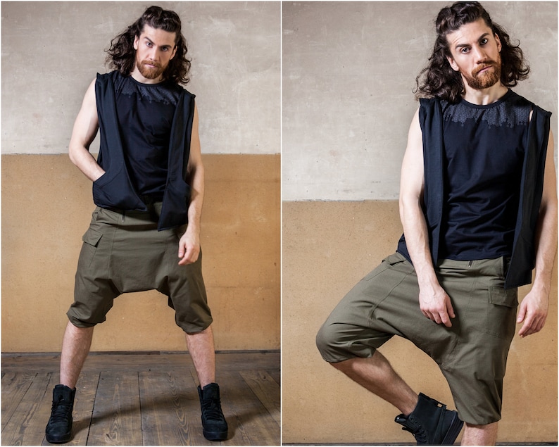 May include: A man wearing a black sleeveless shirt and olive green cargo shorts. He is standing with his left leg bent and his right leg straight. The background is a textured wall.