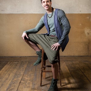 May include: A man wearing a blue vest over a green shirt, green cargo shorts, and brown boots. He is sitting on a wooden stool with a wooden floor behind him.