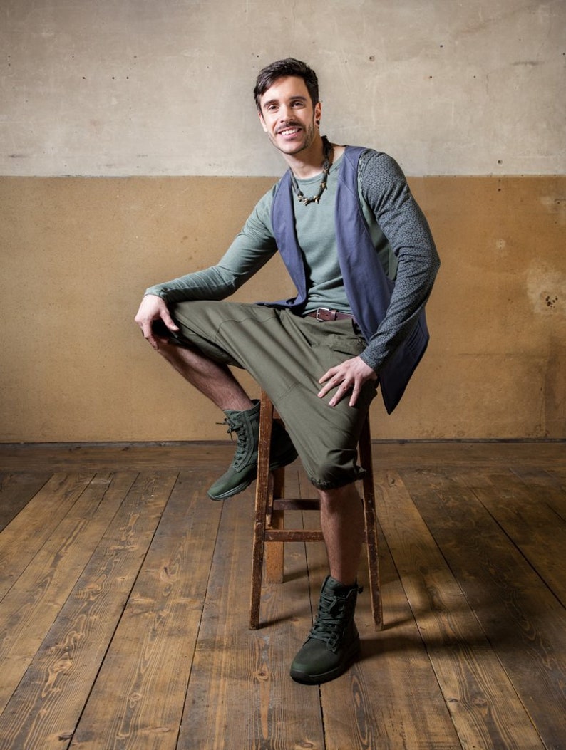 May include: A man wearing a blue vest over a green shirt, green cargo shorts, and brown boots. He is sitting on a wooden stool with a wooden floor behind him.