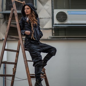 May include: A woman wearing a black hooded jacket and black pants stands on a wooden ladder. She is wearing black boots and has a chain around her waist.
