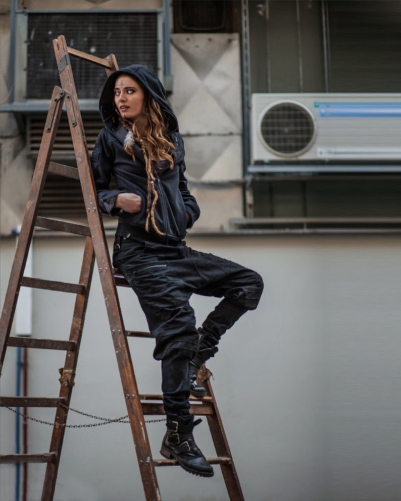 May include: A woman wearing a black hooded jacket and black pants stands on a wooden ladder. She is wearing black boots and has a chain around her waist.