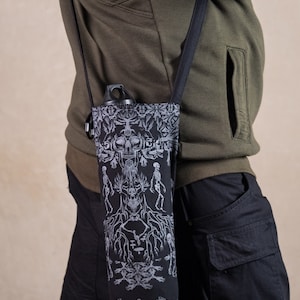 May include: Black water bottle carrier with a black strap, featuring a detailed white and gray design of skulls, skeletons, and tree roots. The carrier is designed to hold a water bottle and is worn over the shoulder.