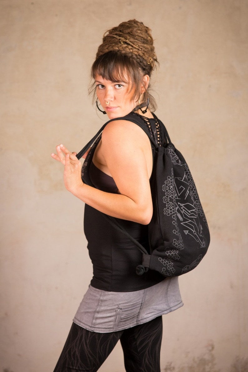 May include: Black drawstring backpack with a geometric design in a lighter color. The bag is worn over the shoulder. The person in the image is wearing a black tank top, gray skirt, and patterned leggings.