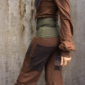 May include: A woman wearing a brown hooded top and brown pants with a green waistband and brown and black patchwork details. The pants have a drawstring at the bottom.