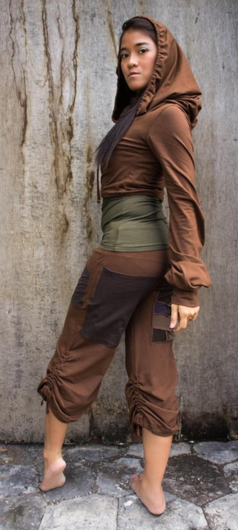 May include: A woman wearing a brown hooded top and brown pants with a green waistband and brown and black patchwork details. The pants have a drawstring at the bottom.