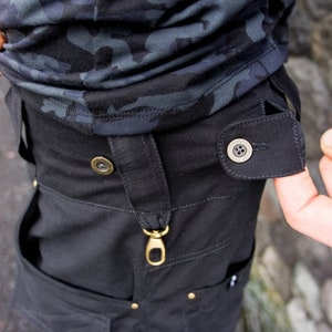 Op de afbeelding: Zwarte werkkleding met een gouden metalen gesp en twee messing knopen. De broek heeft een grote zak aan de voorkant.
