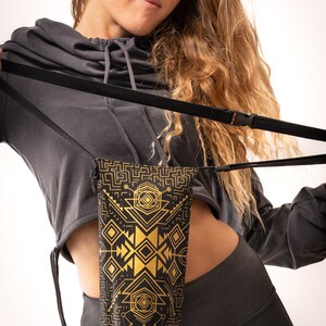 May include: Black and gold geometric pattern phone bag with a black strap. The bag is held by a person wearing a gray hooded sweatshirt and black leggings. The bag's design features gold shapes on a black background.