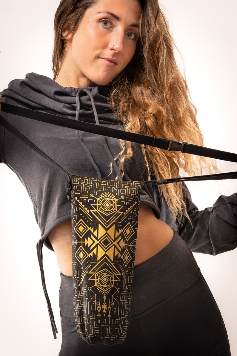 May include: Black and gold geometric pattern phone bag with a black strap. The bag is held by a person wearing a gray hooded sweatshirt and black leggings. The bag's design features gold shapes on a black background.
