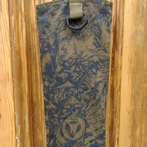 May include: A bottle carrier made of patterned fabric, featuring a green bottle inside. The fabric has a dark olive and black design, with a strap and a metal ring for carrying. The background is a wooden door.