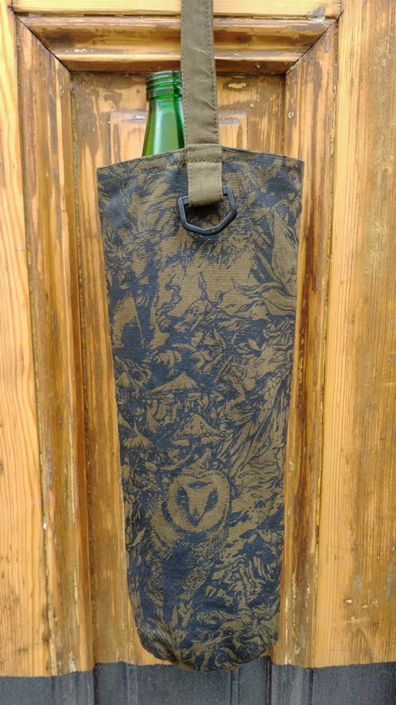 May include: A bottle carrier made of patterned fabric, featuring a green bottle inside. The fabric has a dark olive and black design, with a strap and a metal ring for carrying. The background is a wooden door.