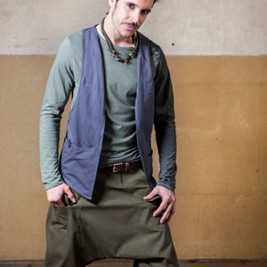 May include: A man wearing a blue vest over a green long-sleeved shirt, brown belt, and green cargo shorts. He is standing in front of a wall with a light brown and gray color scheme.