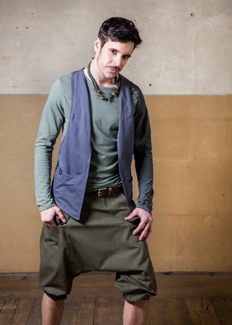 May include: A man wearing a blue vest over a green long-sleeved shirt, brown belt, and green cargo shorts. He is standing in front of a wall with a light brown and gray color scheme.