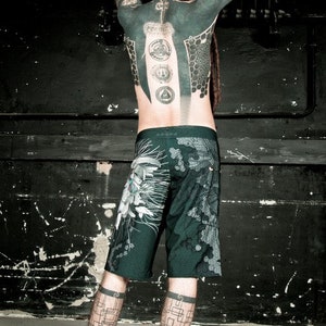 May include: A person with a full body tattoo, including geometric designs and symbols, wearing dark green shorts with a floral pattern. The person has dreadlocks and is standing in front of a black wall.