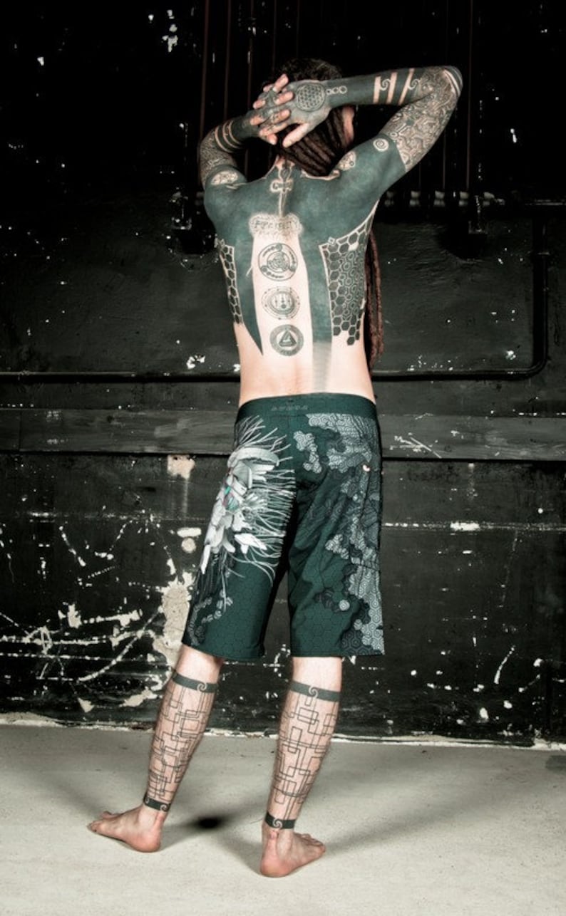 May include: A person with a full body tattoo, including geometric designs and symbols, wearing dark green shorts with a floral pattern. The person has dreadlocks and is standing in front of a black wall.
