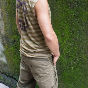 May include: A man wearing a striped, sleeveless shirt and khaki cargo pants stands in front of a mossy wall.