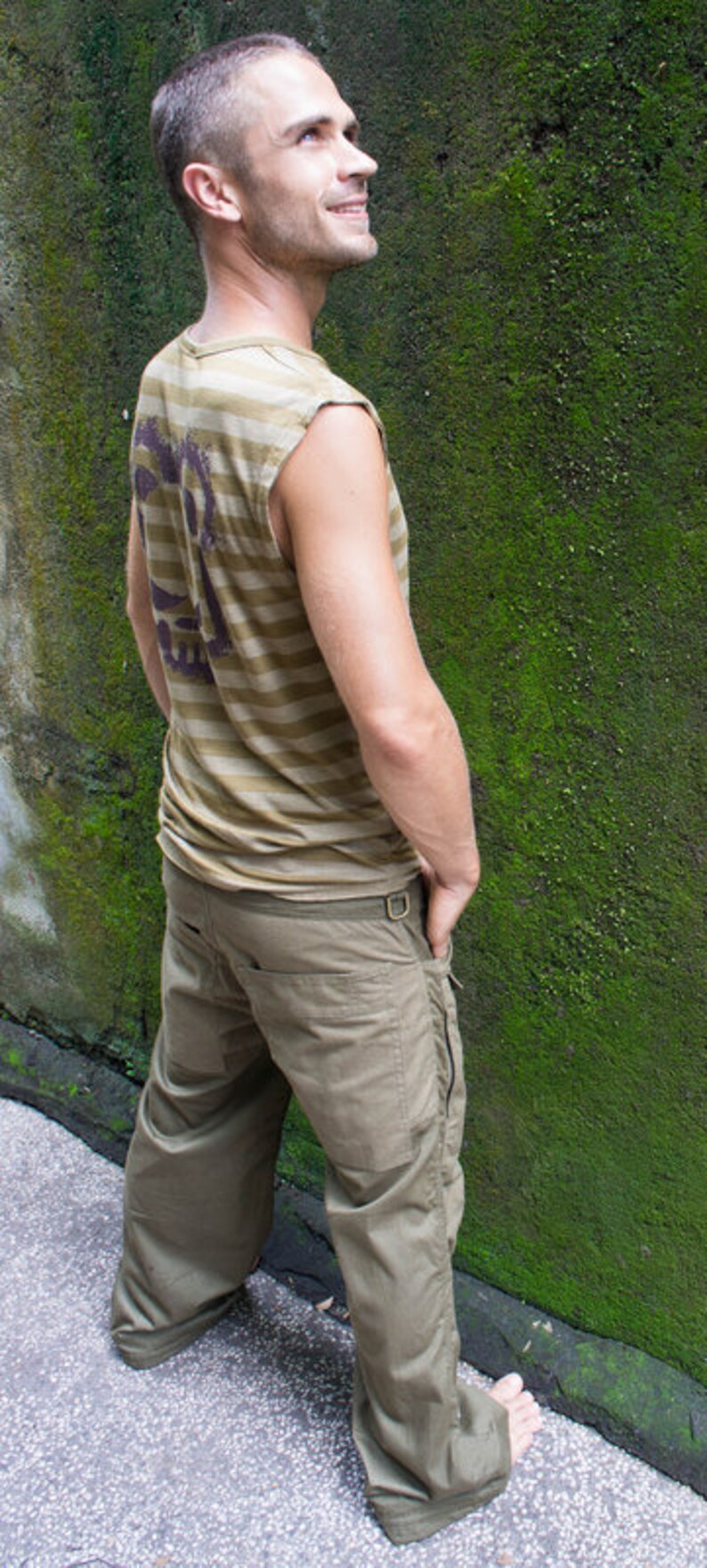 May include: A man wearing a striped, sleeveless shirt and khaki cargo pants stands in front of a mossy wall.