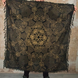 May include: A black and gold geometric patterned throw blanket with fringed edges. The blanket is draped over a person's shoulders.