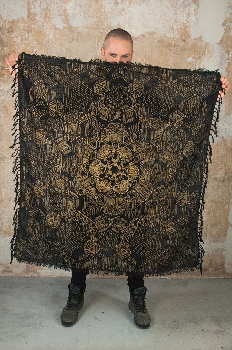 May include: A black and gold geometric patterned throw blanket with fringed edges. The blanket is draped over a person's shoulders.