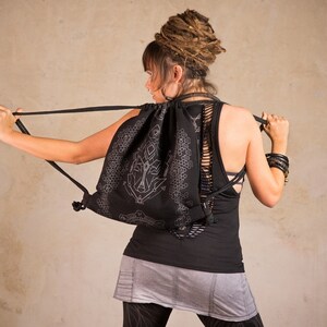May include: Black drawstring backpack with a white geometric design. The backpack has a unique cut-out design on the back. The person is wearing a black tank top and gray skirt. The background is a neutral color.