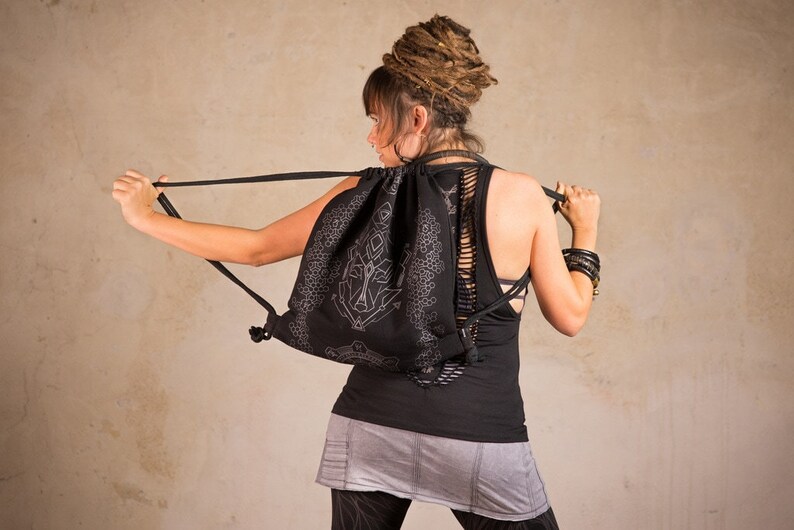 May include: Black drawstring backpack with a white geometric design. The backpack has a unique cut-out design on the back. The person is wearing a black tank top and gray skirt. The background is a neutral color.