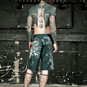 May include: A person with long dreadlocks and full body tattoos is wearing dark green shorts with a geometric pattern. The person is standing in front of a black wall with a metal pipe running horizontally across the wall.