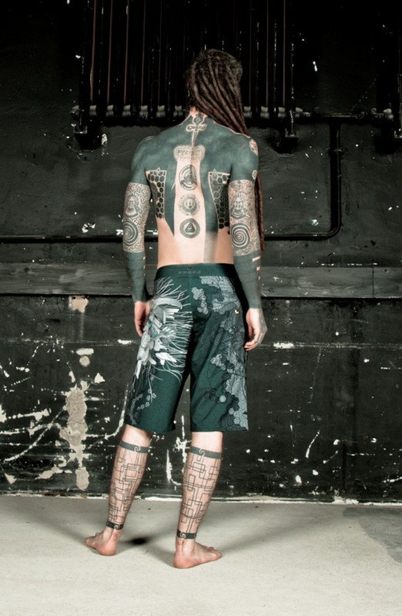 May include: A person with long dreadlocks and full body tattoos is wearing dark green shorts with a geometric pattern. The person is standing in front of a black wall with a metal pipe running horizontally across the wall.