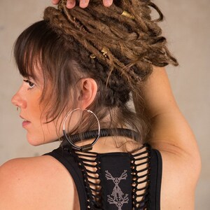 May include: A woman with brown dreadlocks, wearing a black tank top with a unique cut-out design on the back. She has large silver hoop earrings and a black choker. Her hair is styled with gold-colored hair accessories.