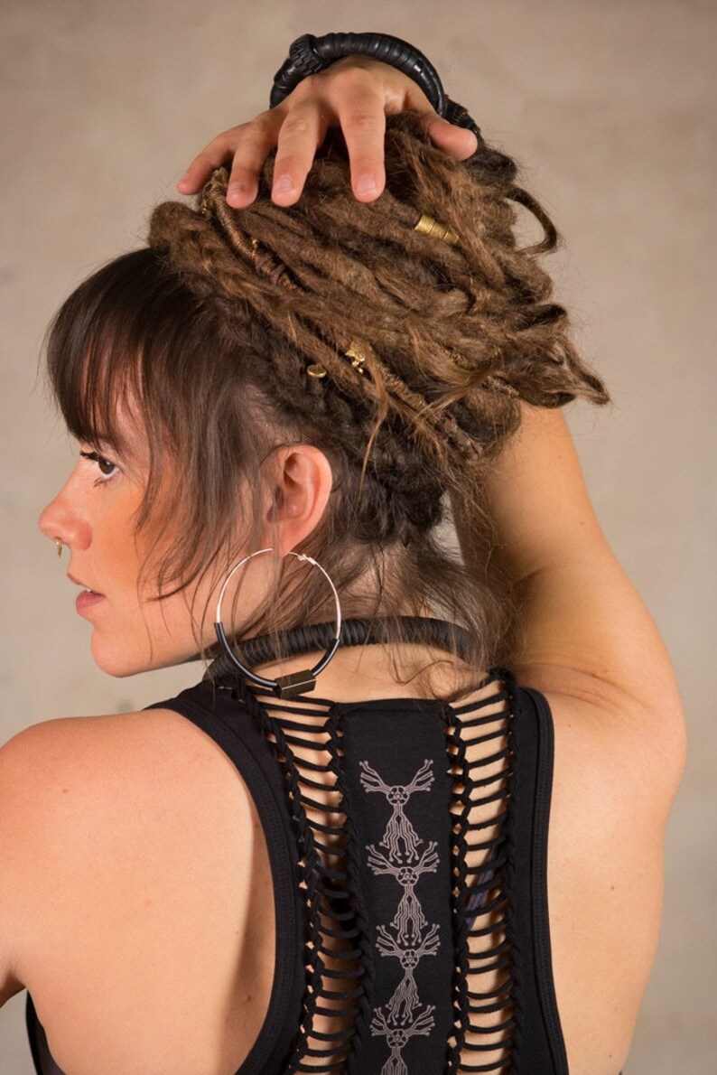 May include: A woman with brown dreadlocks, wearing a black tank top with a unique cut-out design on the back. She has large silver hoop earrings and a black choker. Her hair is styled with gold-colored hair accessories.