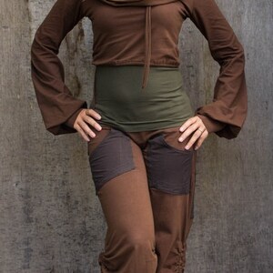 May include: A woman wearing a brown, long-sleeved top with a cowl neck and brown pants with patch pockets. She is standing in front of a wood-grain background.