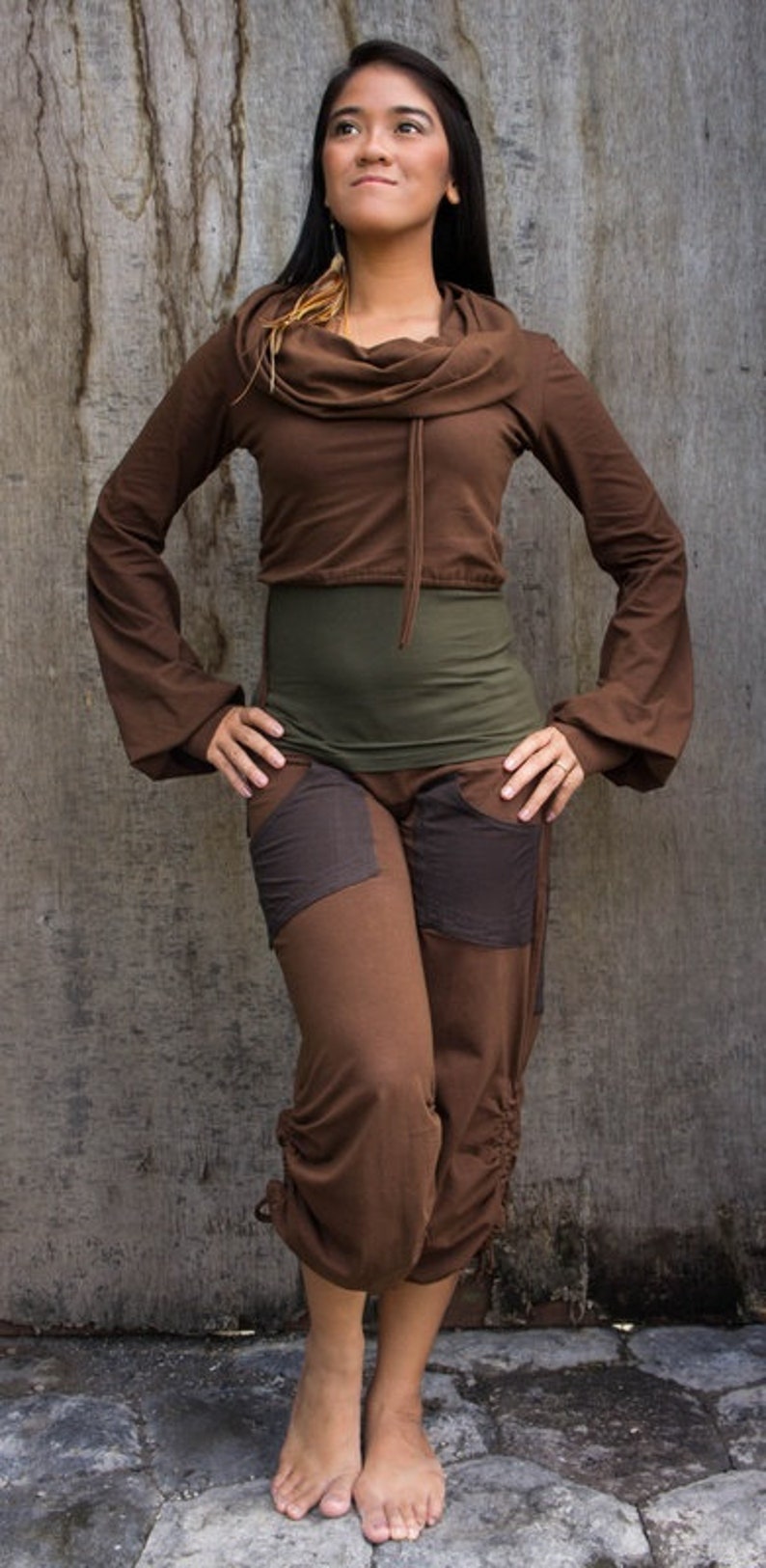 May include: A woman wearing a brown, long-sleeved top with a cowl neck and brown pants with patch pockets. She is standing in front of a wood-grain background.