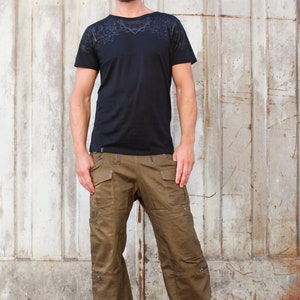 May include: A man wearing a black t-shirt with a geometric pattern on the chest and brown cargo pants. He is standing in front of a metal wall.