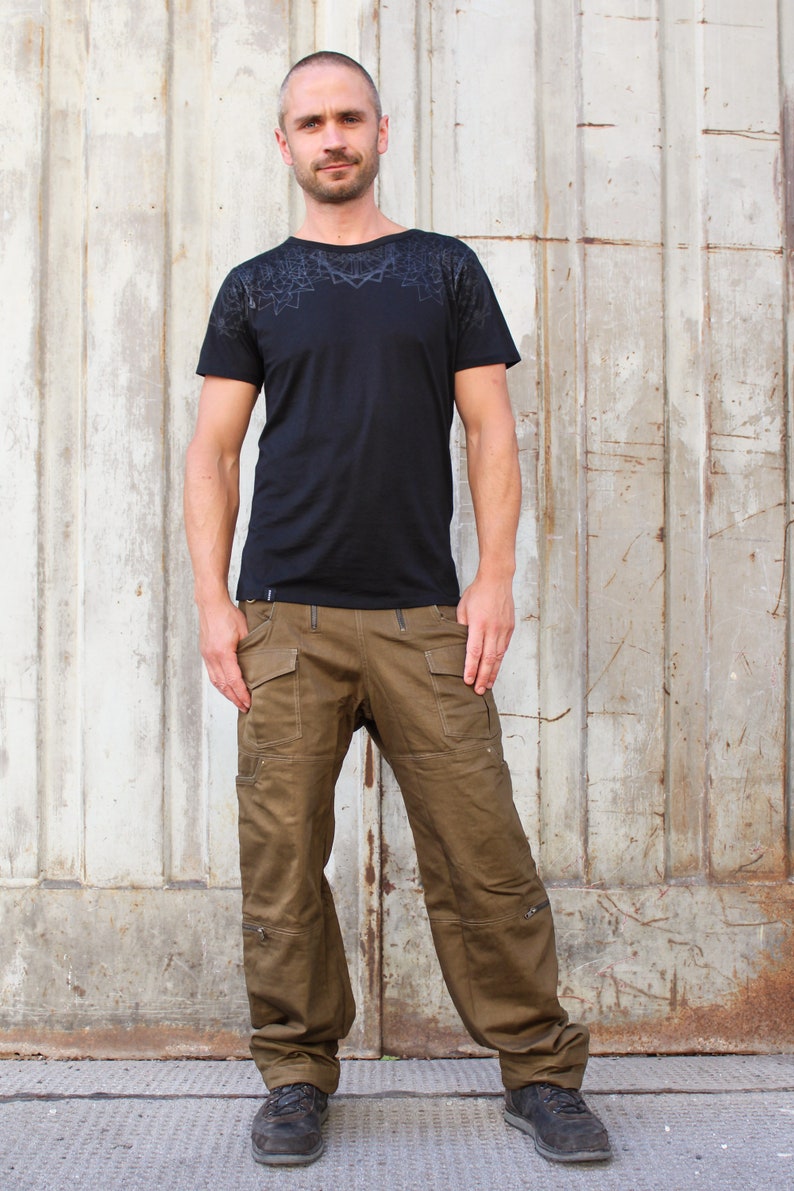 May include: A man wearing a black t-shirt with a geometric pattern on the chest and brown cargo pants. He is standing in front of a metal wall.