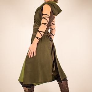 May include: A woman wearing a long, dark green tunic with brown leather straps and boots. The tunic has a hood and is tied at the waist. The woman is standing with her back to the camera and looking over her shoulder.