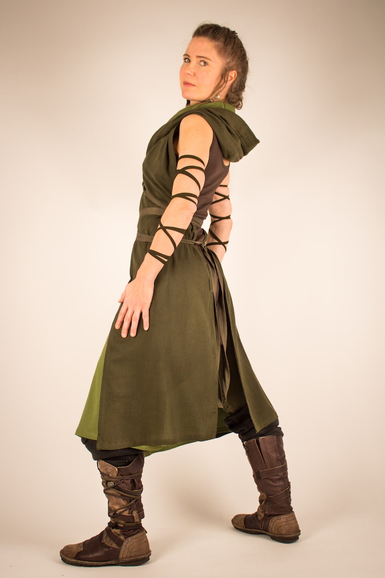 May include: A woman wearing a long, dark green tunic with brown leather straps and boots. The tunic has a hood and is tied at the waist. The woman is standing with her back to the camera and looking over her shoulder.