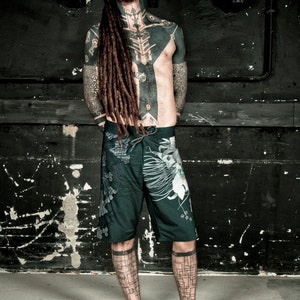 May include: A man with long dreadlocks and extensive black tattoos is wearing dark green and white patterned shorts. He is standing in front of a dark, textured wall.