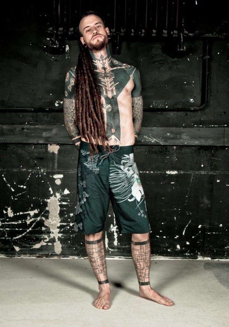 May include: A man with long dreadlocks and extensive black tattoos is wearing dark green and white patterned shorts. He is standing in front of a dark, textured wall.