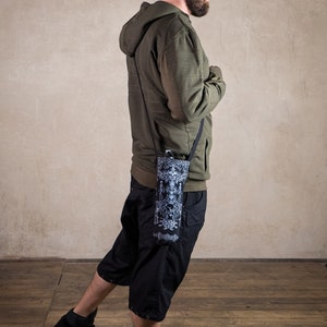 May include: A person wearing a green hooded sweatshirt, black shorts, and black sneakers. A black and white patterned water bottle holder is slung across the body. The holder has a black strap.
