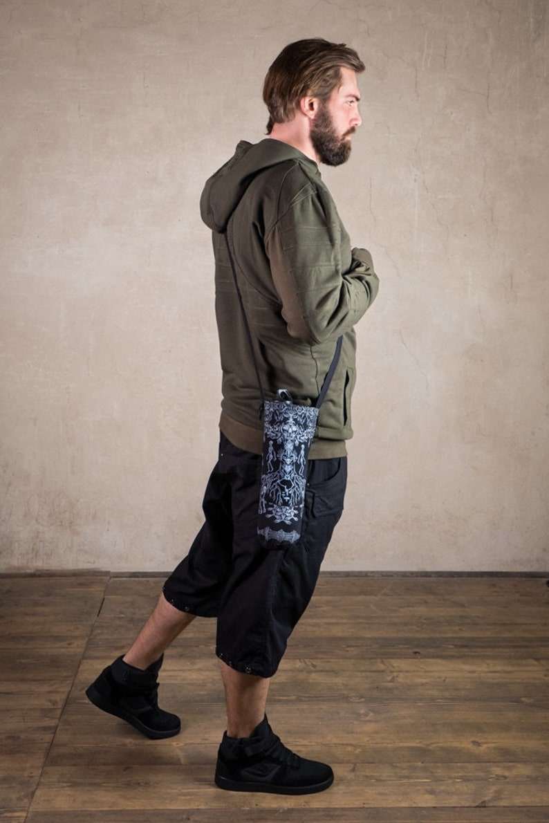 May include: A person wearing a green hooded sweatshirt, black shorts, and black sneakers. A black and white patterned water bottle holder is slung across the body. The holder has a black strap.