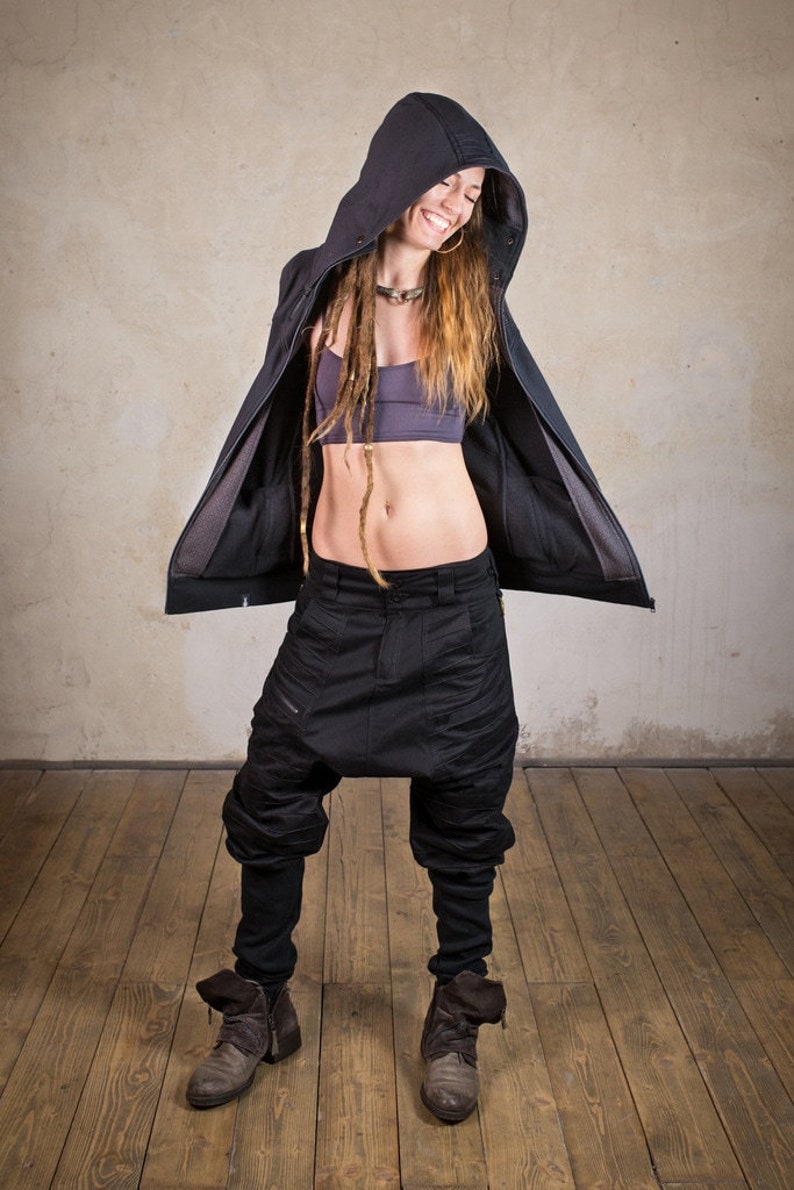 May include: A woman wearing a black hooded jacket with the hood up, black pants, and brown boots. She has long blonde dreadlocks and is smiling.