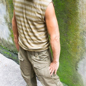 May include: A man wearing a striped, sleeveless shirt and khaki cargo pants. He is standing in front of a wall covered in moss.