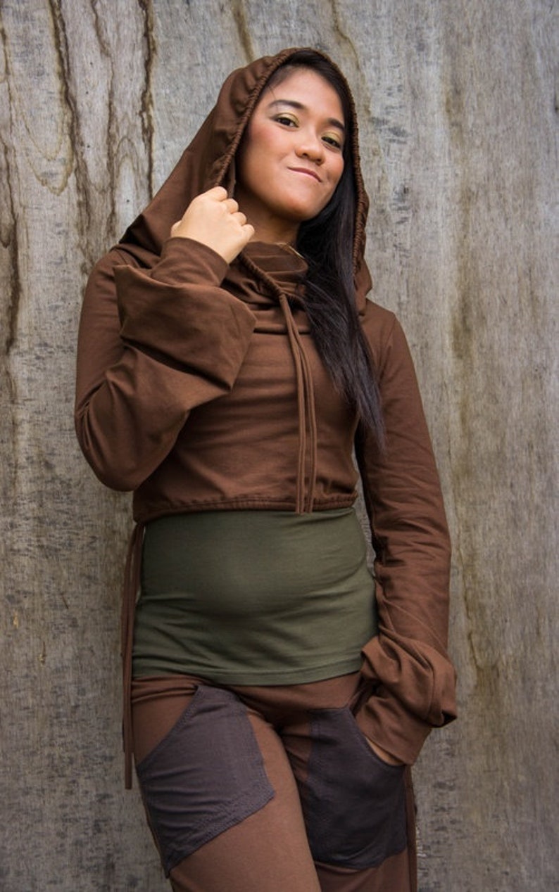 May include: A brown hooded top with long sleeves and a drawstring at the neck. The top is cropped and has a relaxed fit. The model is wearing a green tank top underneath and brown pants with pockets.