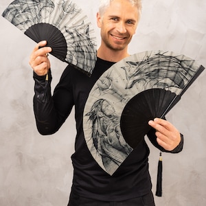 May include: A person holding two black and white hand fans with intricate designs. The fans are made of paper and have black handles with tassels. The designs on the fans feature a woman, a wolf, and a moon.
