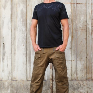 May include: A man wearing a black t-shirt with a geometric pattern and brown cargo pants. He is standing in front of a metal wall.